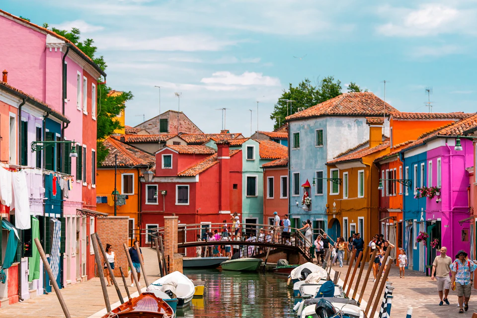 A canal in a colorful town with people on a bridge, surrounded by boats and vibrant buildings under a clear sky.