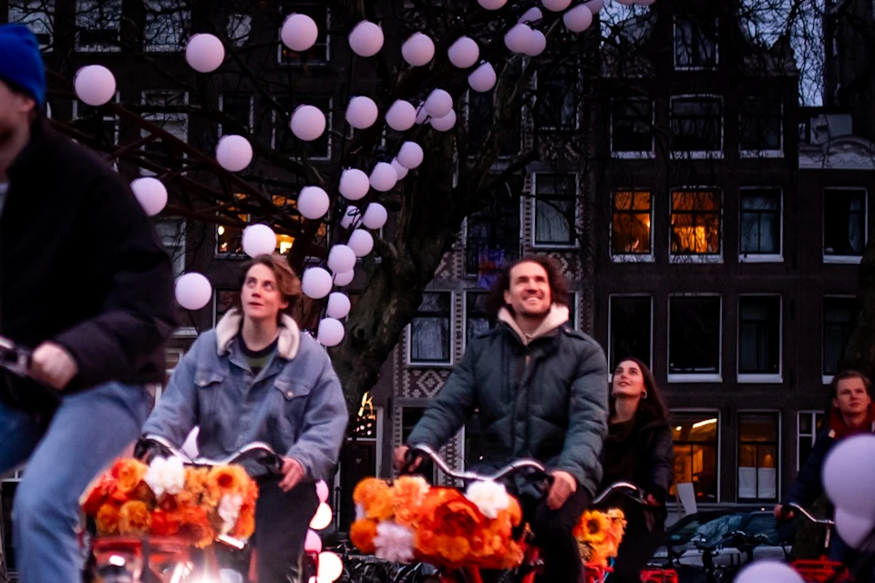 Three people riding bikes decorated with orange flowers under a tree adorned with hanging white lights in an urban setting.