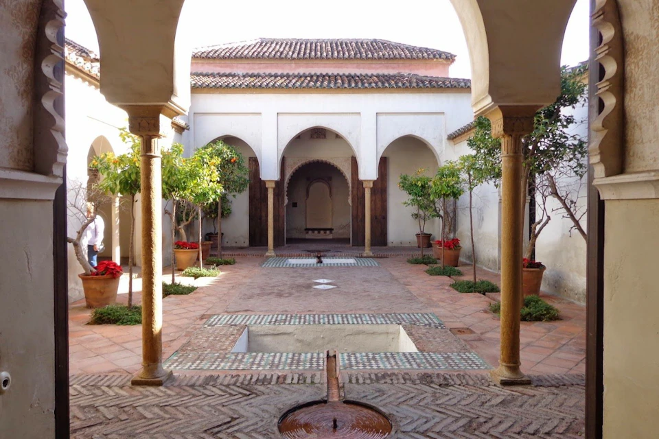A courtyard with a central water feature, surrounded by arches, potted trees, and a tiled walkway under a tiled roof structure.
