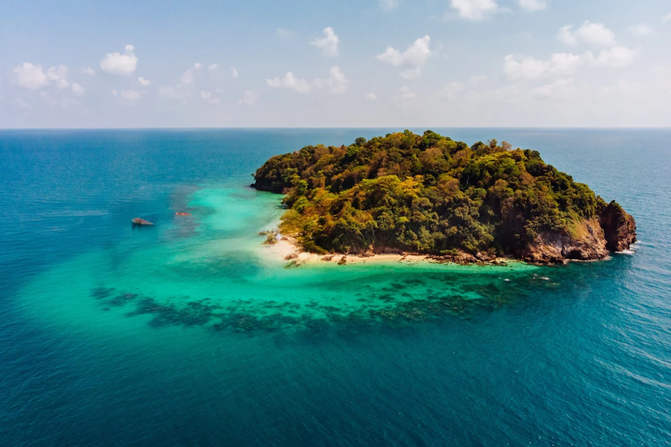 Aerial view of a small, lush island surrounded by turquoise water, with a sandy beach and clear blue sky.