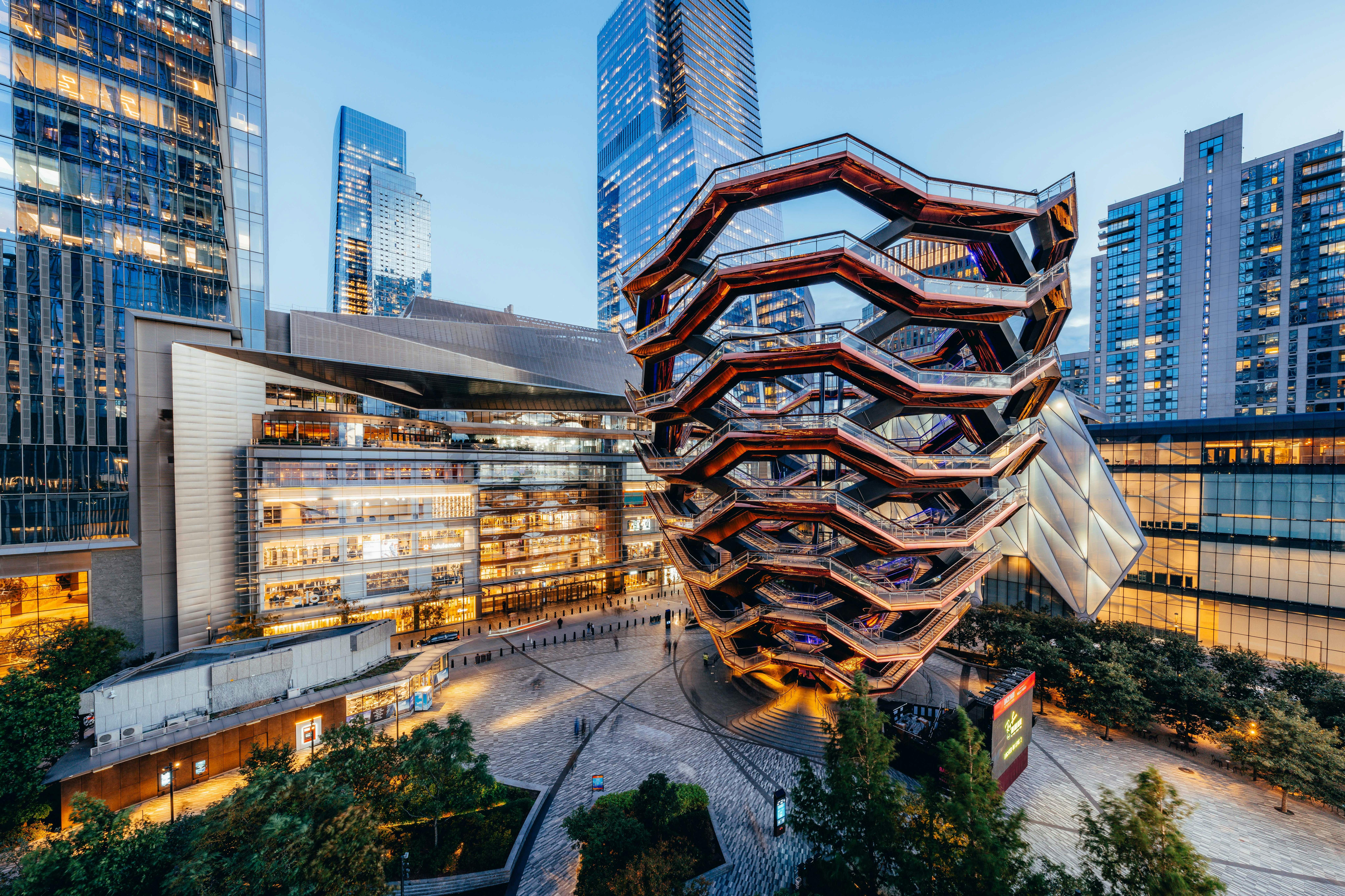 Futuristic multi-level structure with interconnected stairs, surrounded by tall modern buildings and a plaza with greenery.