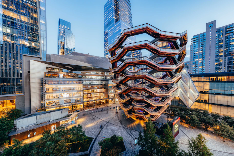 Futuristic multi-level structure with interconnected stairs, surrounded by tall modern buildings and a plaza with greenery.