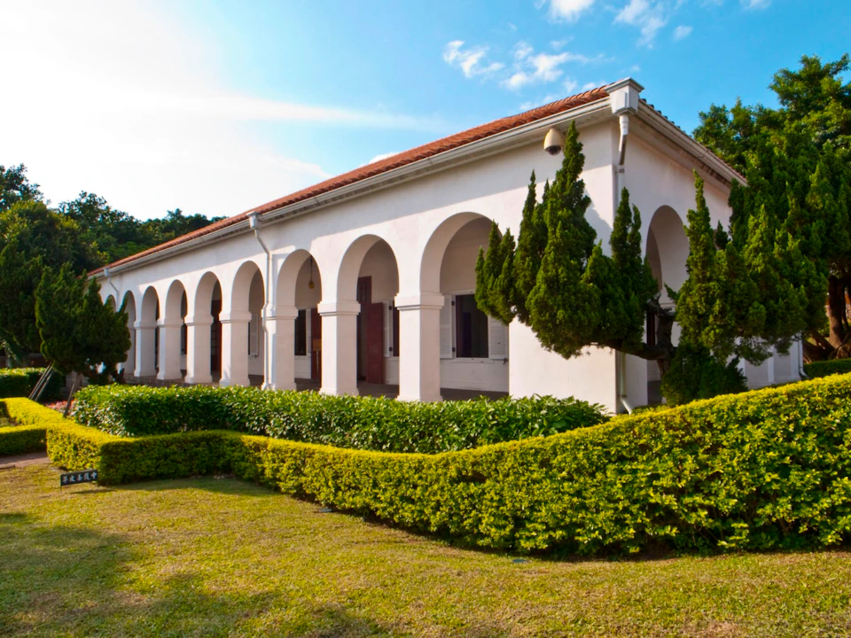 A white building with arched columns, surrounded by trimmed shrubs and trees, under a blue sky with light clouds.