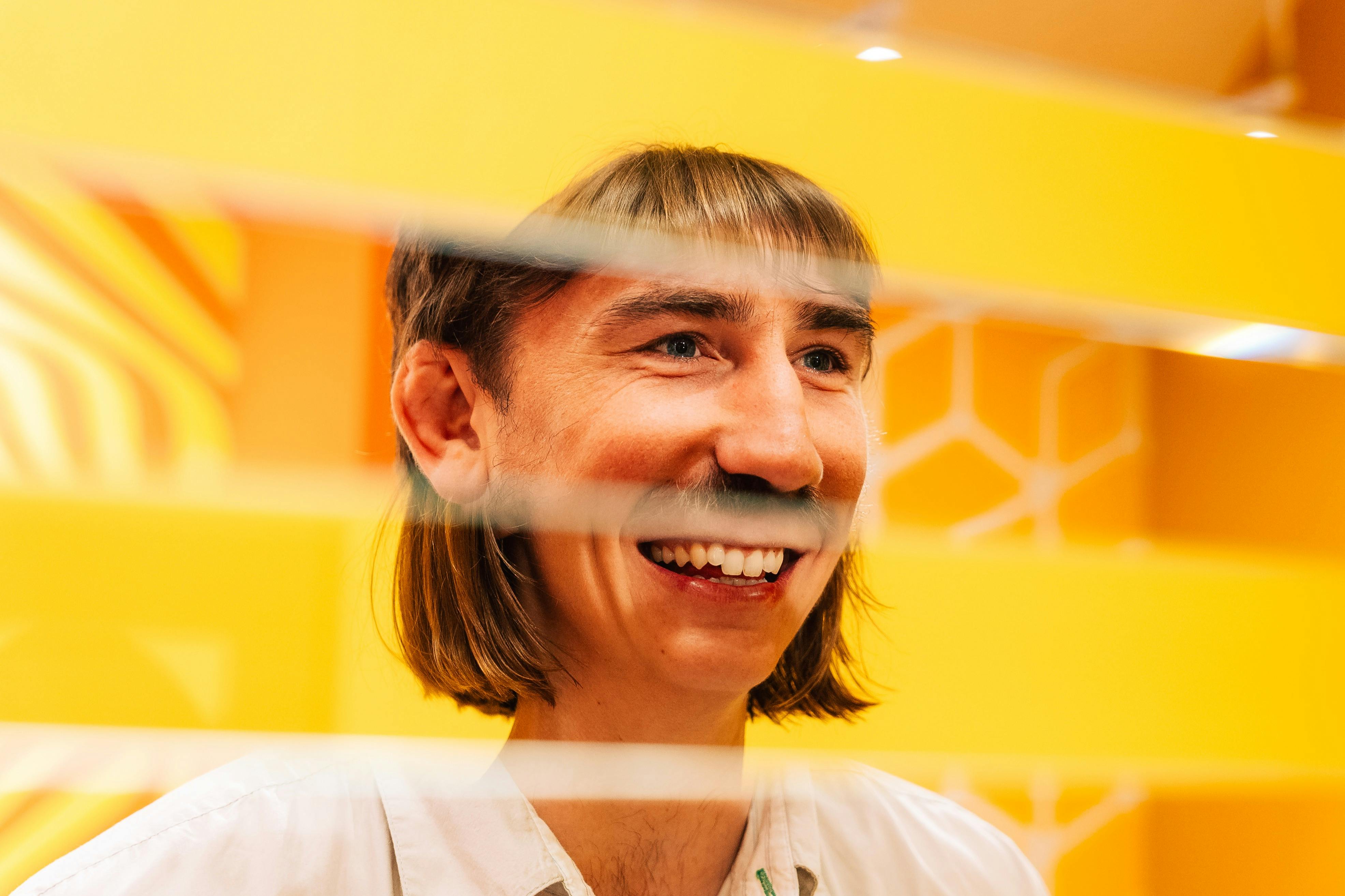 A smiling person with shoulder-length hair is seen through horizontal blinds against a bright yellow background with hexagon patterns.