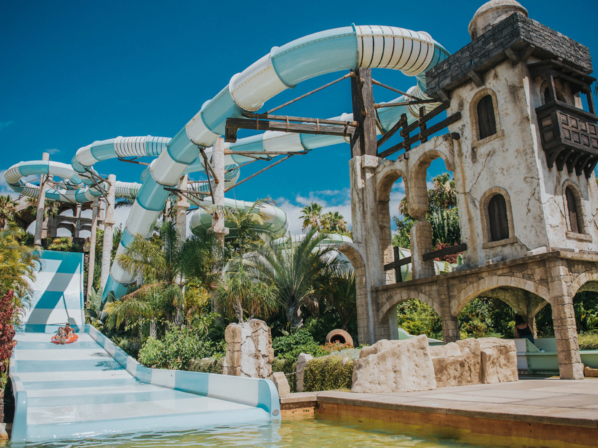 Outdoor water park with large blue and white tube slides, surrounded by palm trees and a stone structure, under a clear blue sky.
