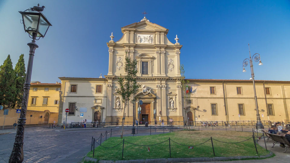 A historic, ornate church façade with statues and decorative elements under a clear blue sky, surrounded by a small fenced grassy area.