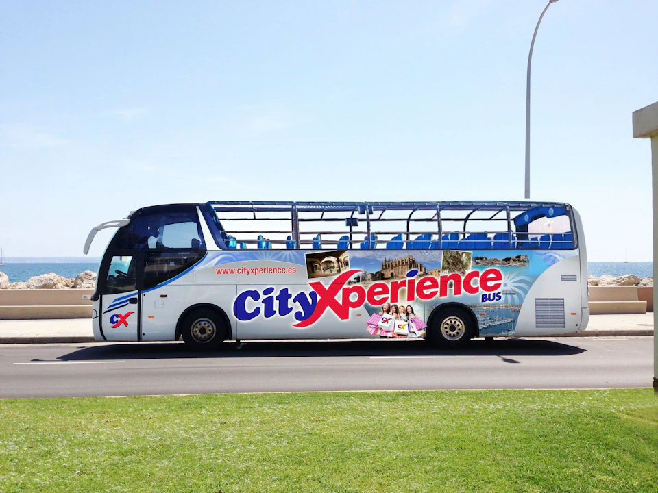 A white double-decker City Xperience bus with an open top and colorful graphics is parked on a road beside a grassy area.