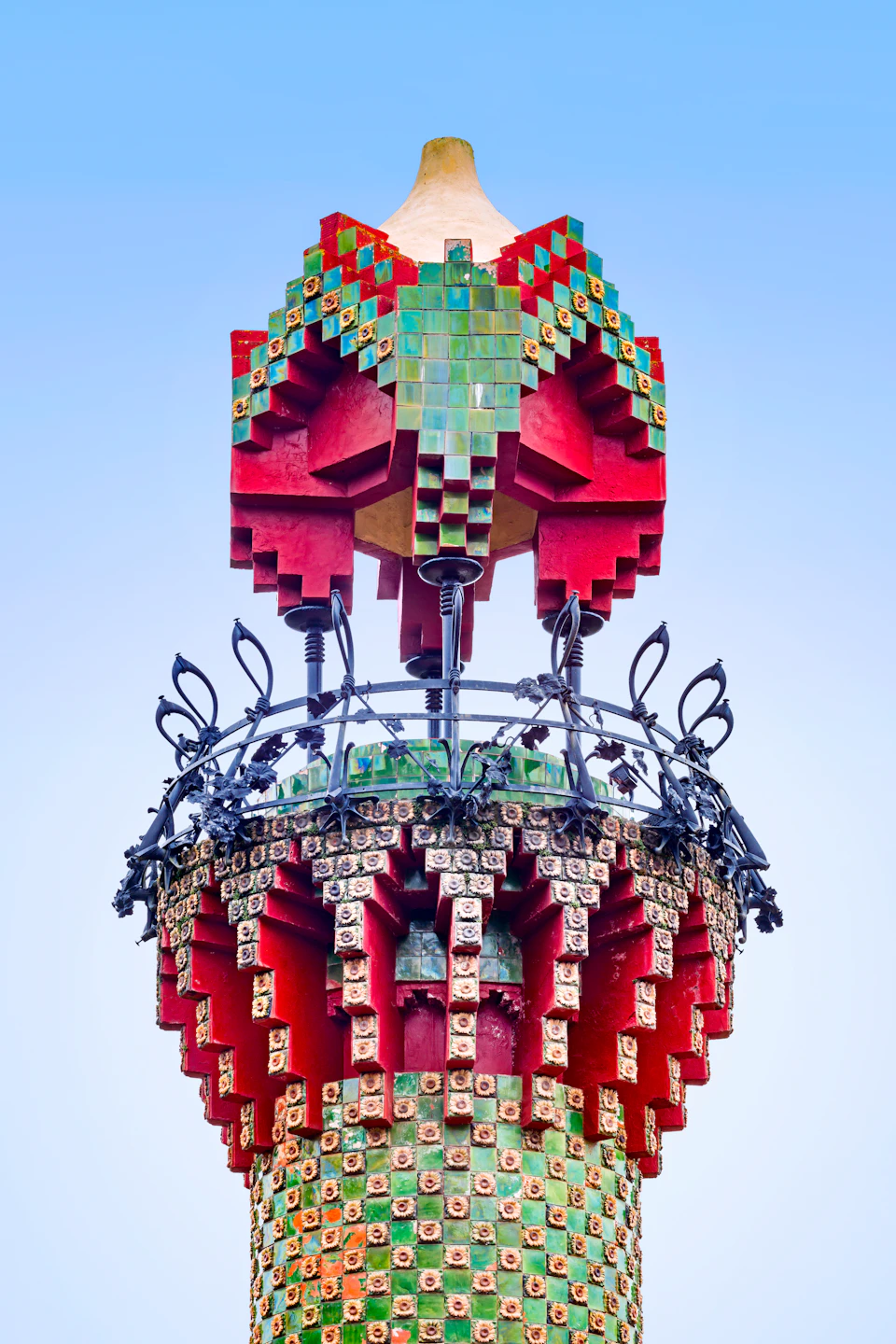 Ornate tower top with red and green geometric mosaic patterns, black wrought iron railing, and blue sky background.