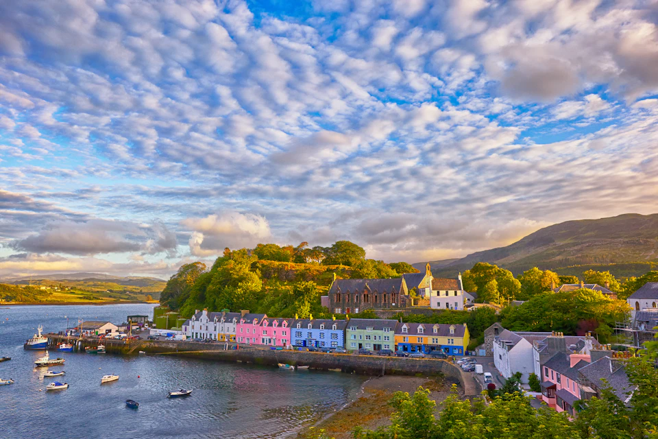 A row of colorful houses along a harbor with boats, lush greenery, and a partly cloudy sky above.