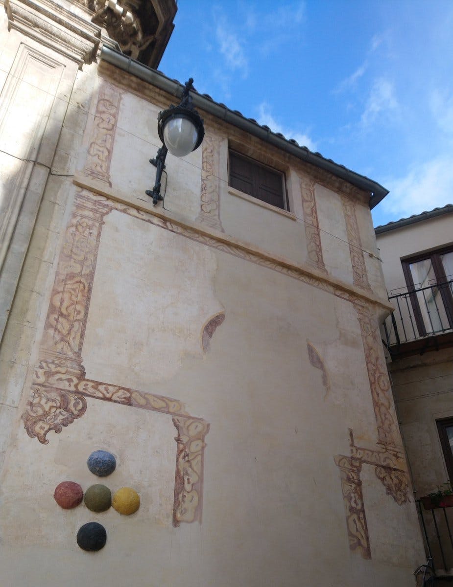 Ein hohes beigefarbenes Gebäude mit dekorativen Verzierungen, einer Straßenlaterne und farbigen Kugeln an der Wand. Der Himmel ist teilweise sichtbar.