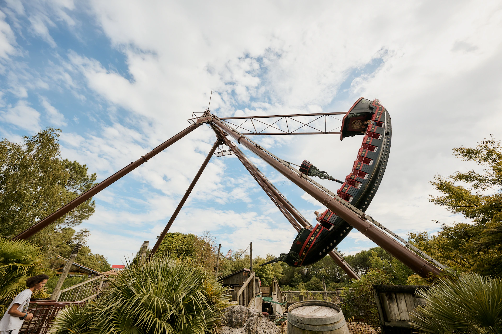 A large pirate ship ride swings high in the air at an amusement park, surrounded by trees and a partly cloudy sky.
