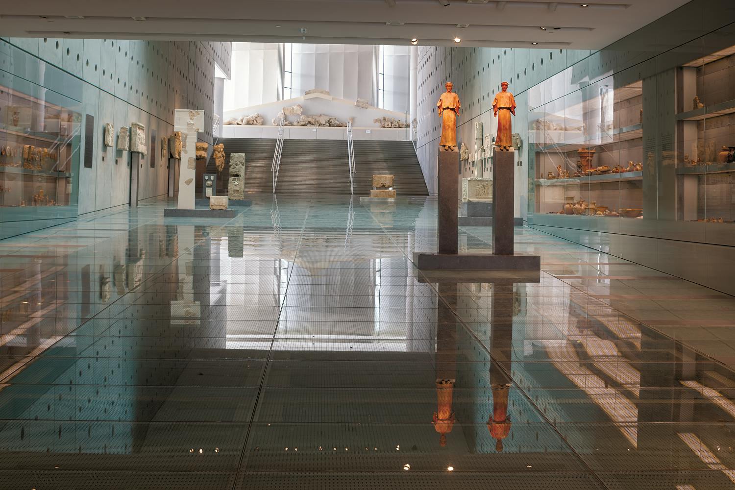 Интерьер музея со стеклянным полом, каменными артефактами и большими статуями. Лестница ведет на верхний уровень с большим количеством отображаемых предметов.