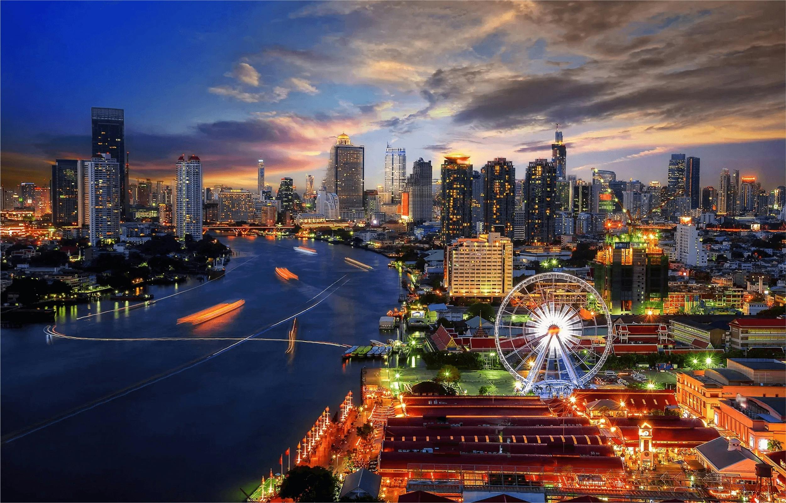 Cityscape at dusk with a river, illuminated buildings, a ferris wheel, and boats leaving light trails on the water.