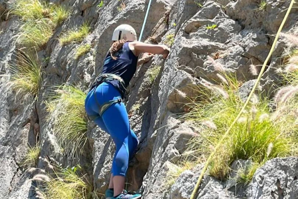 A person wearing a helmet and climbing gear ascends a rocky cliff face surrounded by patches of grass.