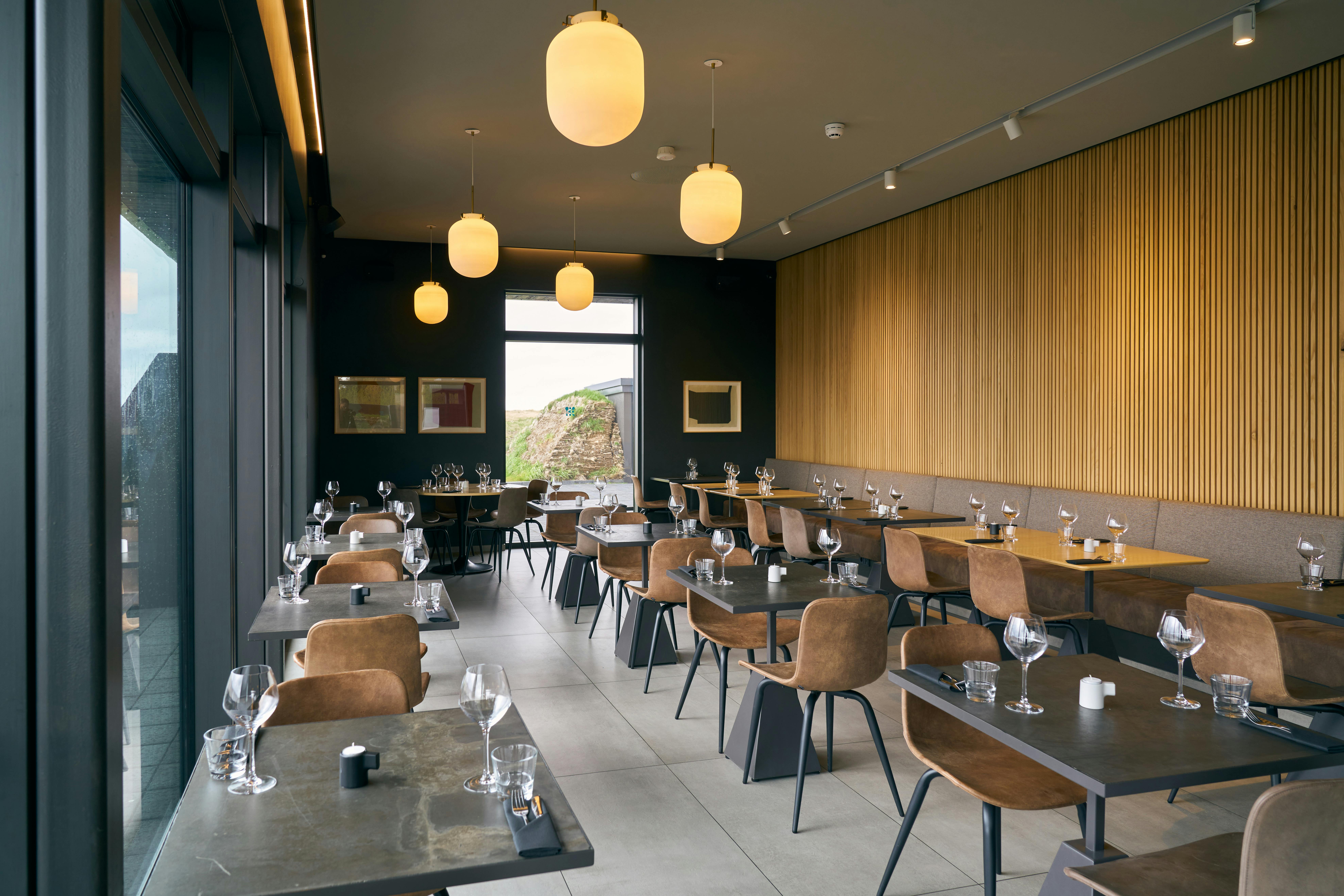 Μοντέρνο εστιατόριο με καφέ καρέκλες, τραπέζια με ποτήρια κρασιού, μεγάλα παράθυρα, τοίχους με ξύλινη επένδυση και κρεμαστά φώτα υδρογείου.