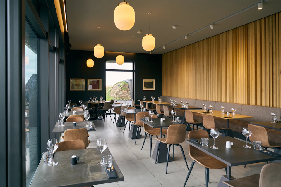 Modern restaurant with brown chairs, set tables with wine glasses, large windows, wood-paneled walls, and hanging globe lights.