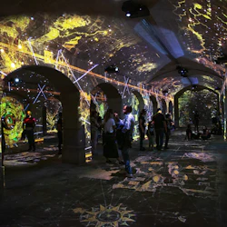 Människor i ett välvt rum med upplysta, färgglada projektioner på valv, tak och golv, som visar invecklade mönster och kartor.