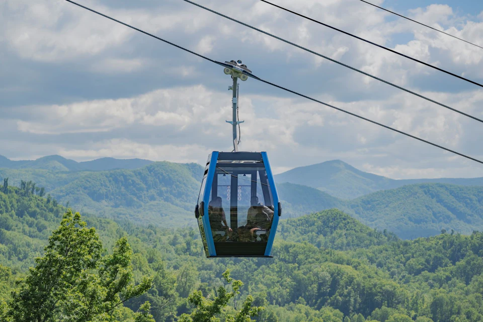 A blue gondola lift suspended over a lush green forest, with mountains and a partly cloudy sky in the background.