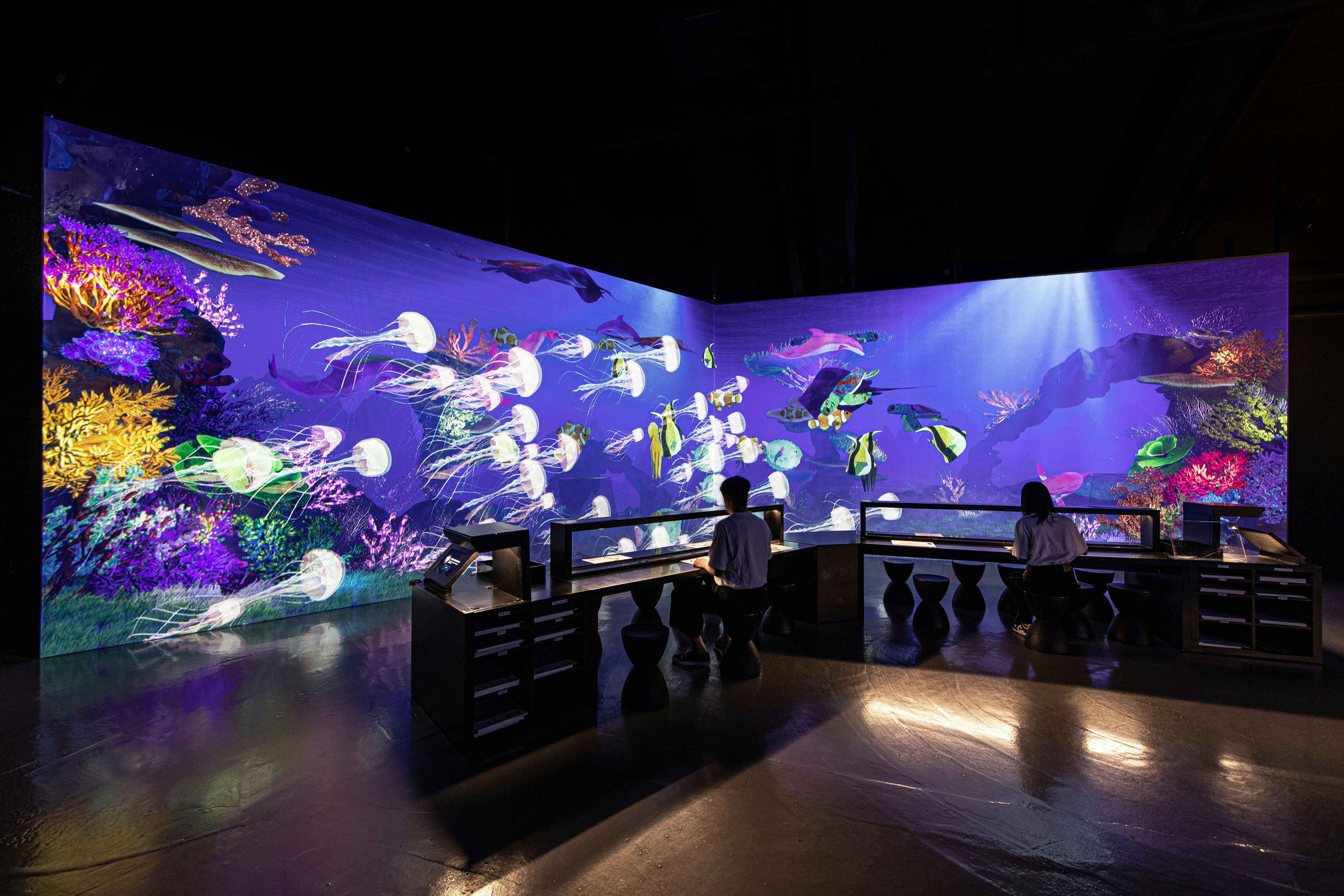 Dwie osoby siedzą przy biurkach przed dużym, wciągającym cyfrowym wyświetlaczem akwarium z meduzami i różnymi kolorowymi rybami.