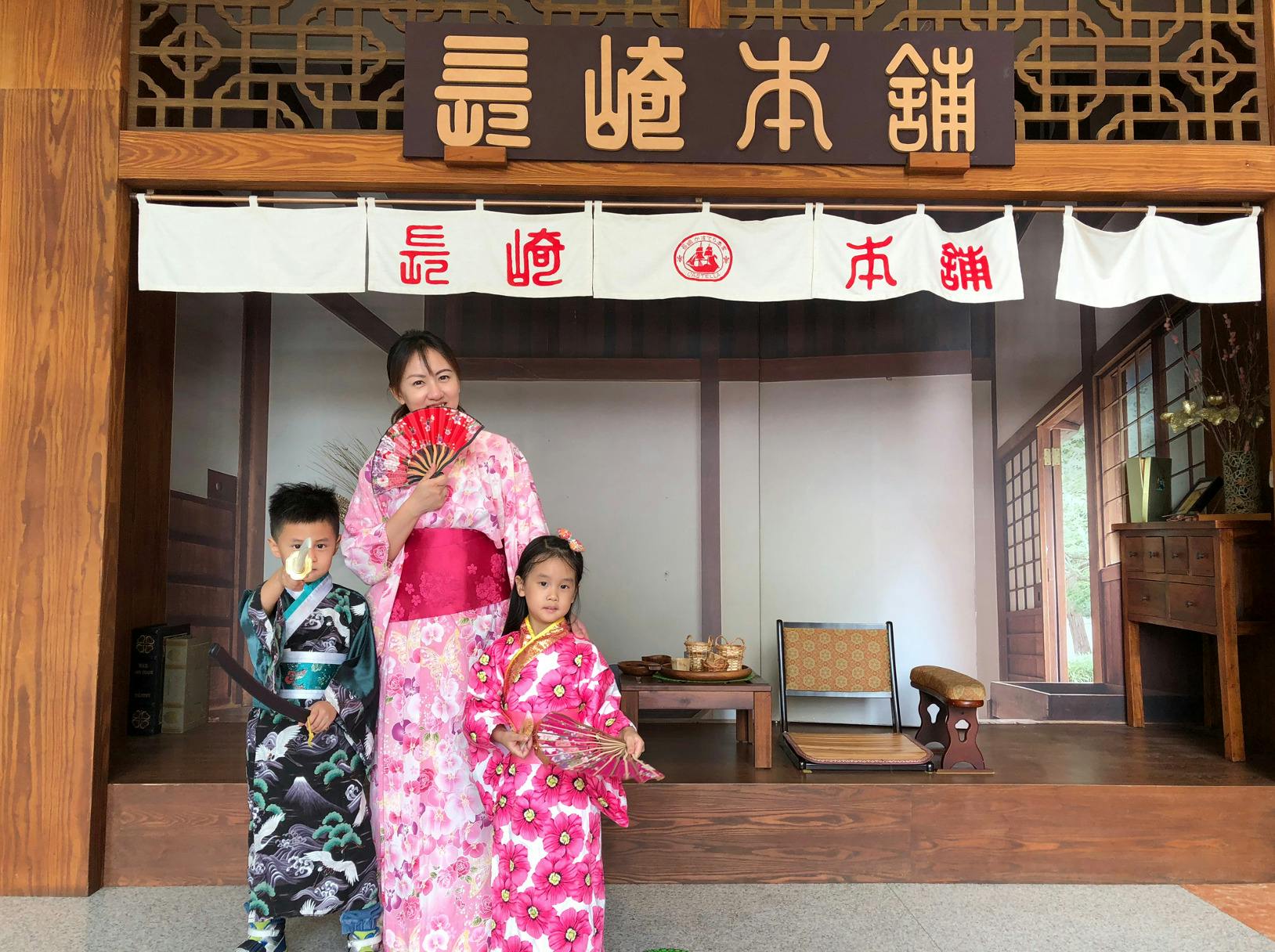 A woman and two children in traditional attire pose in front of a wooden building with a Japanese-style design.