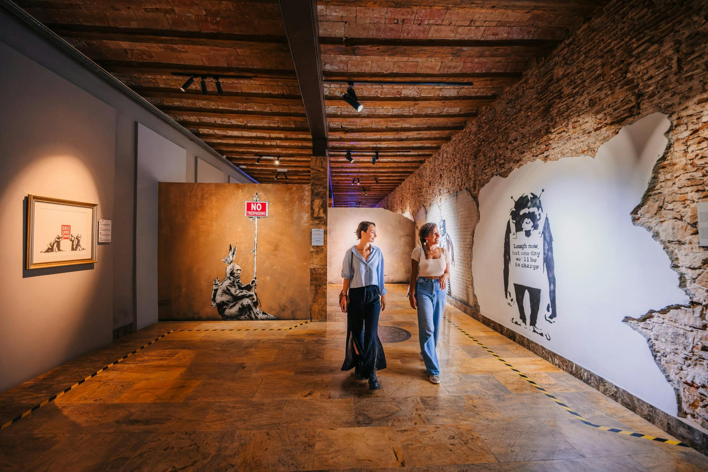Deux personnes traversent une galerie d'art avec des encadrements et des peintures murales sous des poutres en briques apparentes et des projecteurs.