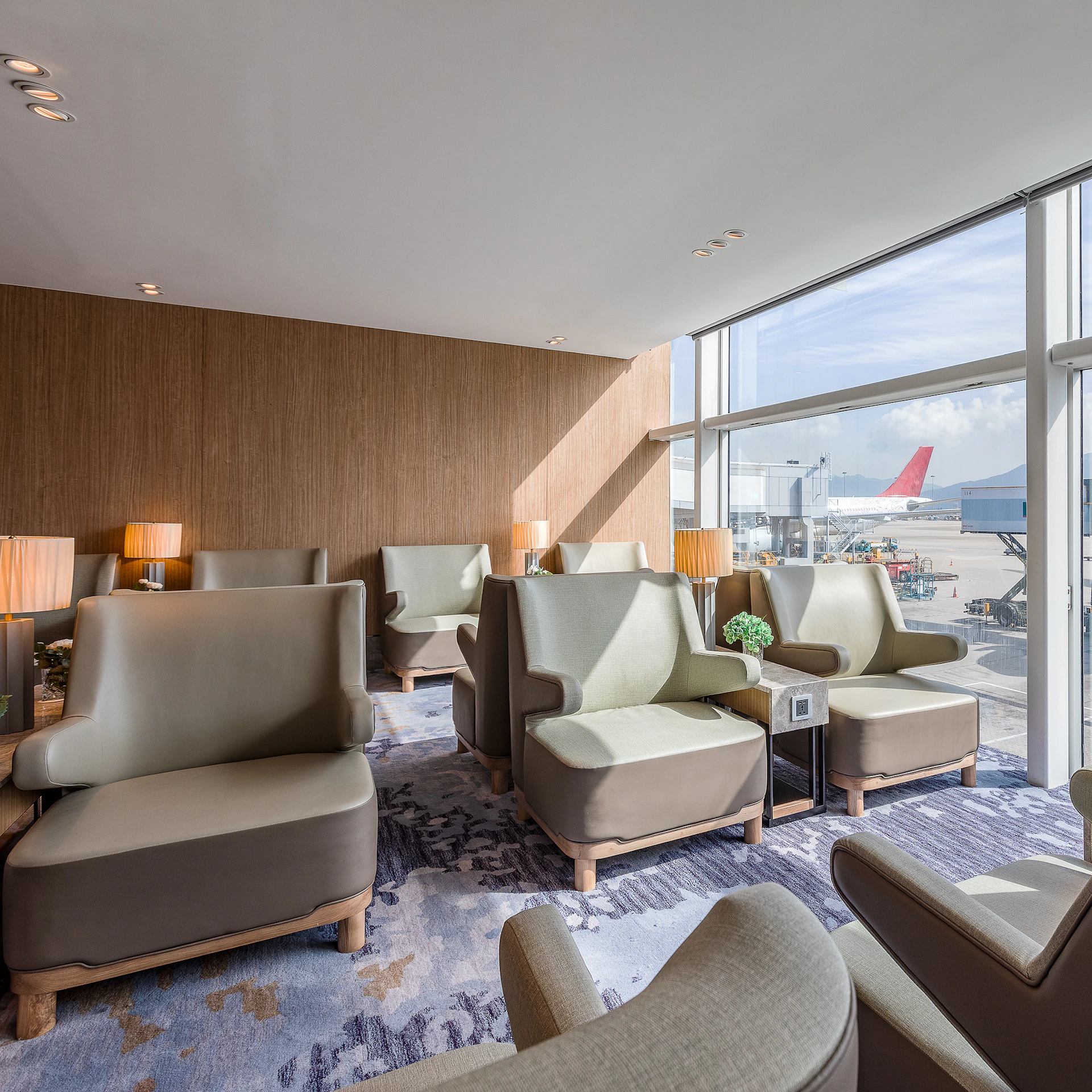 Luxurious airport lounge with beige armchairs, small tables, and lamps. Large windows reveal parked airplanes outside.