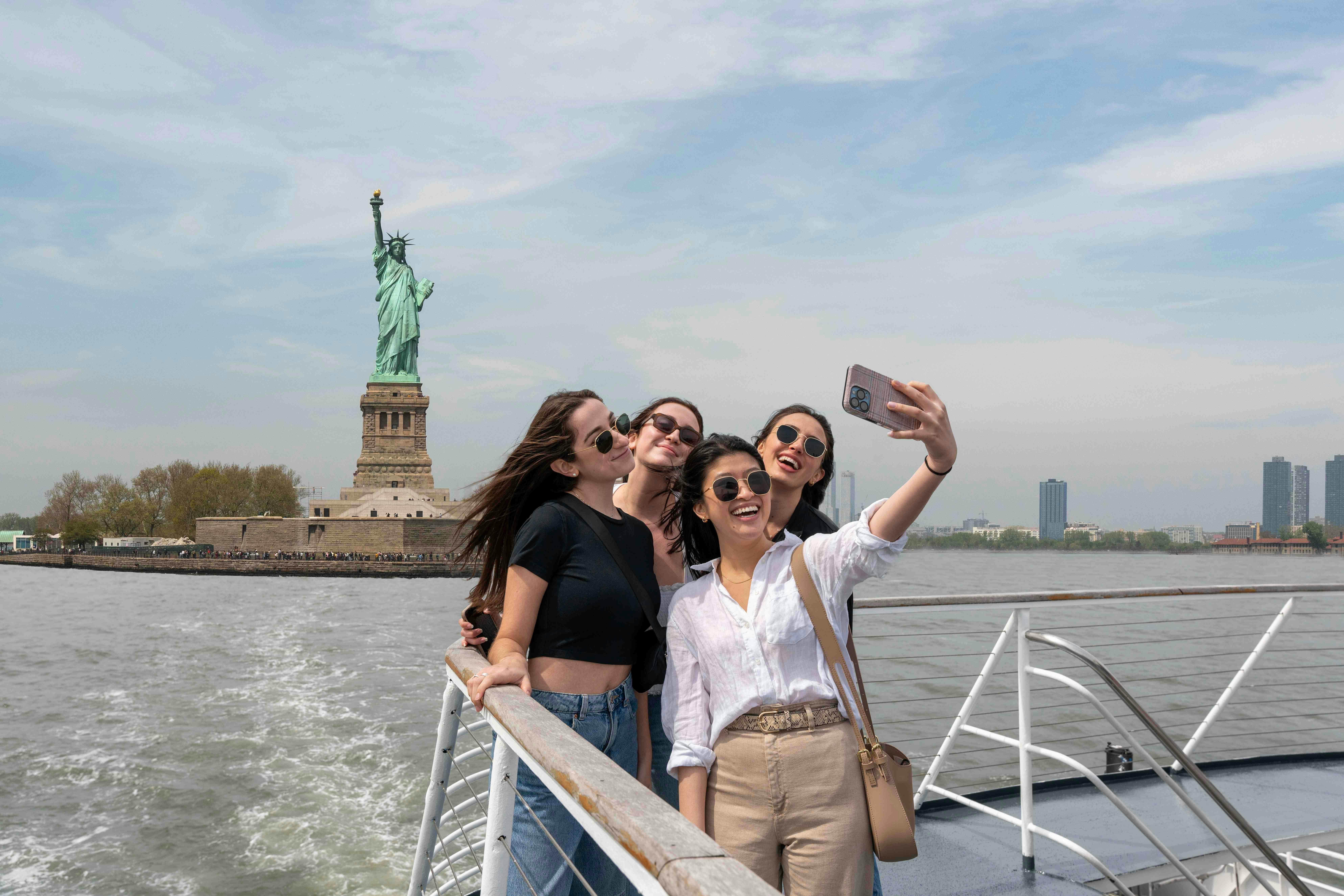 Fire venner tager en selfie på en båd med Frihedsgudinden synlig i baggrunden på en klar dag.