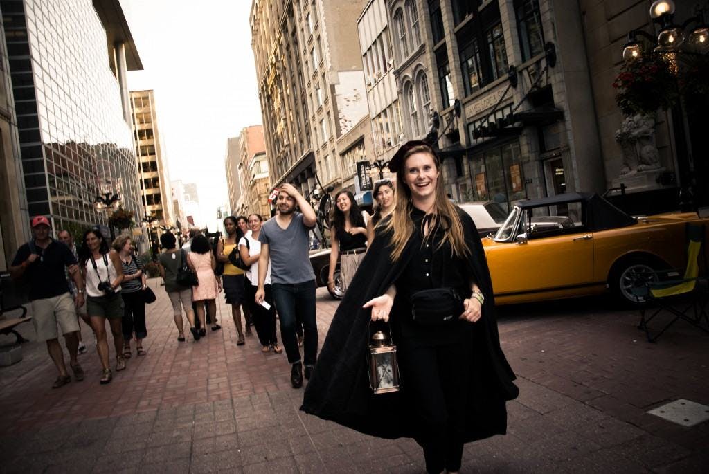 身穿黑色斗篷的微笑女士带着灯笼走在繁华的城市街道上，街道两旁布满了各种各样的人和高楼大厦。