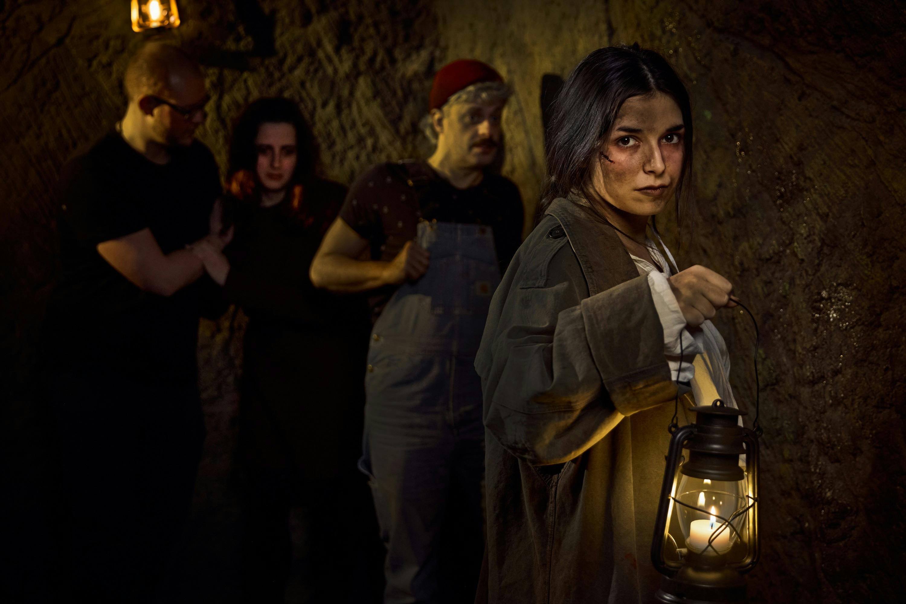 En kvinde, der holder en lanterne i en svagt oplyst hule, med en mand iført overalls og en rød hat i baggrunden.