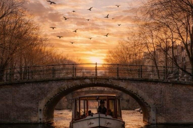 Una barca che passa sotto un ponte in mattoni su un canale al tramonto, con gli uccelli che volano nel cielo e gli alberi che costeggiano il corso d'acqua.