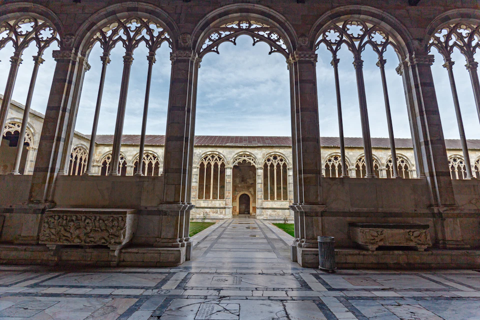 Medieval courtyard with arched stone windows, intricate carvings, and a tiled pathway, framed by gothic columns.