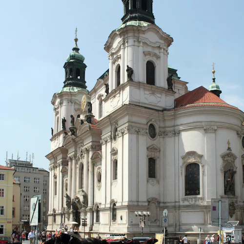 Historisk byggnad med utsmyckad vit fasad och grönklädda torn. Hästdragna vagnar och fotgängare i förgrunden.