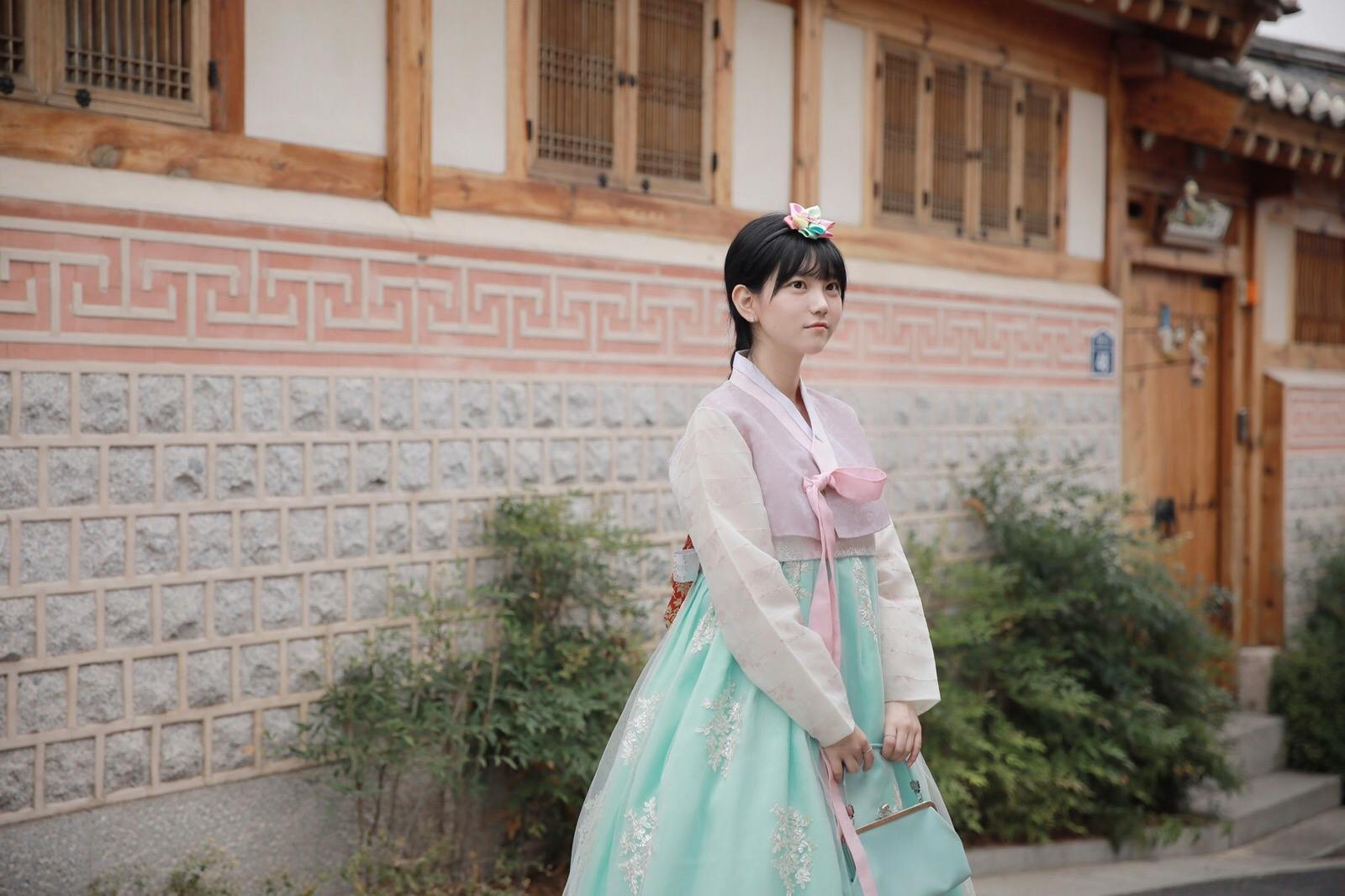 A person wearing traditional Korean clothing stands beside a traditional building with wooden and stone elements.