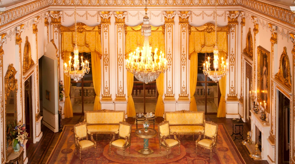 A luxurious room with ornate furniture, golden drapes, and chandeliers. A table with a tea set is in the center.