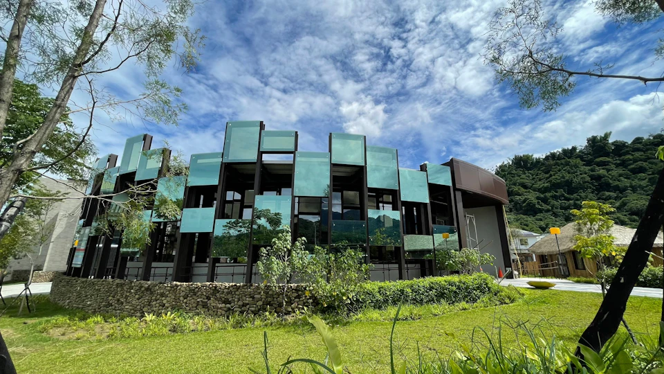 Modern building with reflective glass panels, surrounded by greenery, under a partly cloudy blue sky.