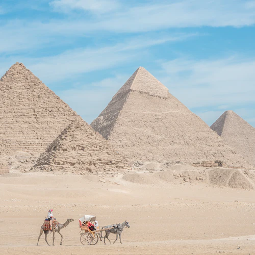 Drei große Pyramiden in der Wüste bei strahlend blauem Himmel, im Vordergrund ein Kamelgespann, das Menschen transportiert.