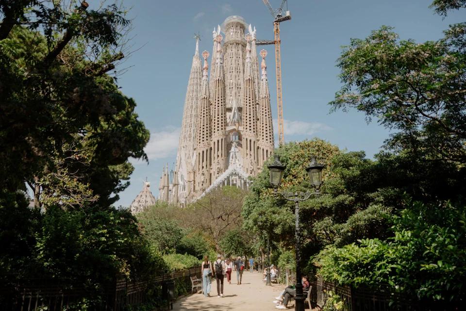A view of the Sagrada Família basilica in Barcelona, surrounded by greenery and people walking along a park pathway.