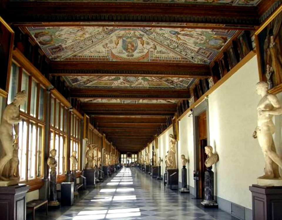 Long corridor with ornate painted ceiling, lined with statues on pedestals, large windows on one side, and a checkered floor.