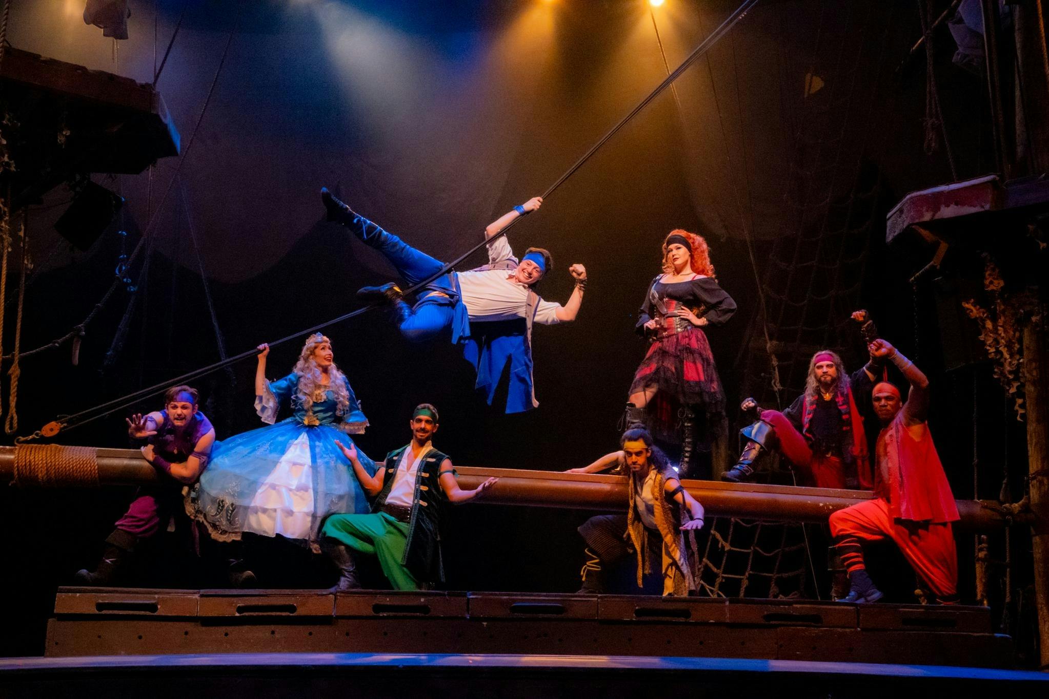 Les artistes, vêtus de costumes colorés, agissent sur une scène conçue comme un navire, avec des gréements et des éclairages spectaculaires.