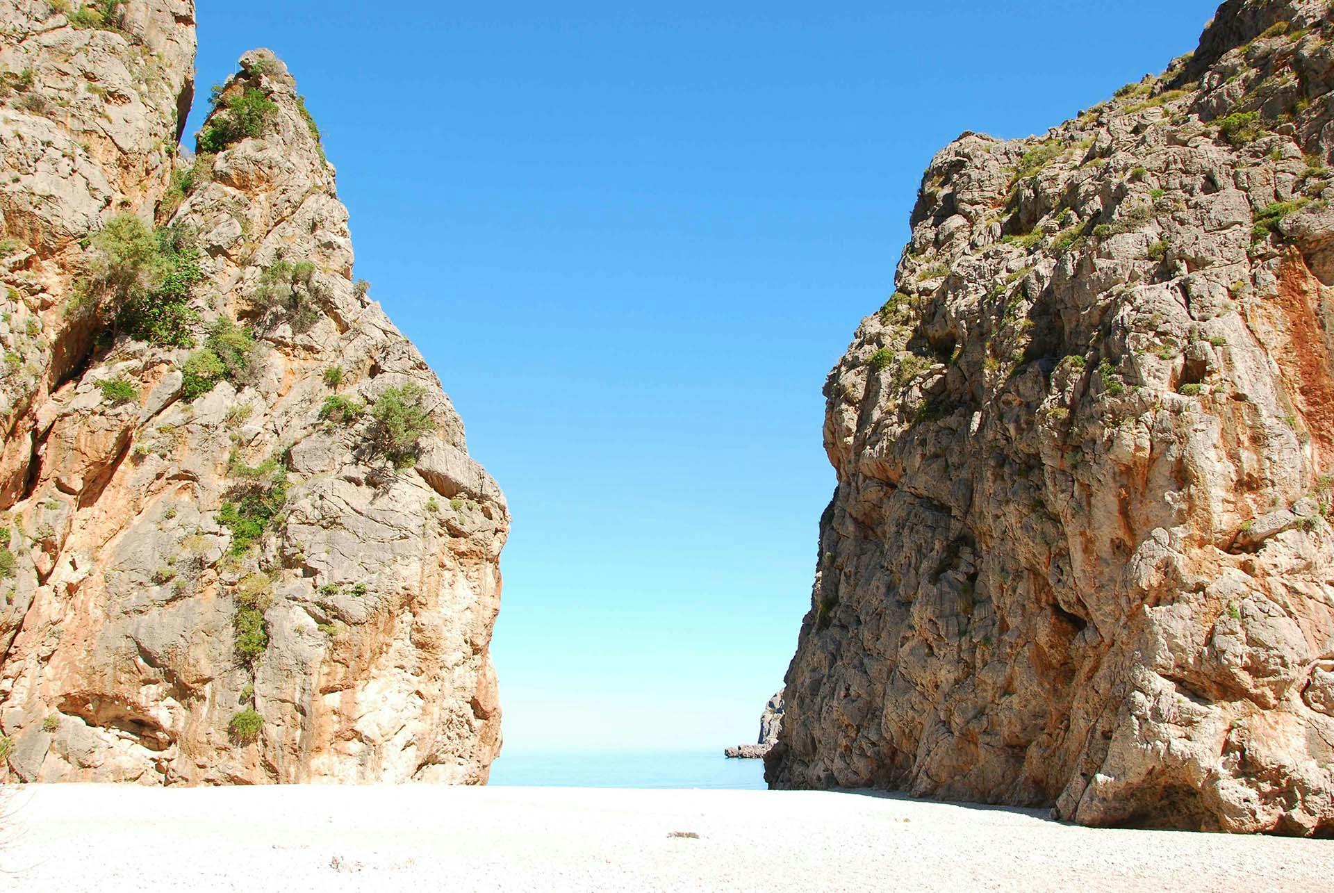 A narrow beach flanked by two large rocky cliffs, with a clear blue sky and calm sea in the background.
