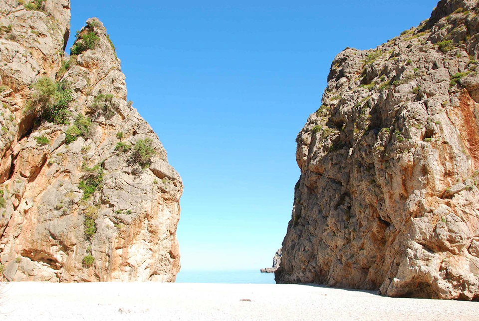 A narrow beach flanked by two large rocky cliffs, with a clear blue sky and calm sea in the background.