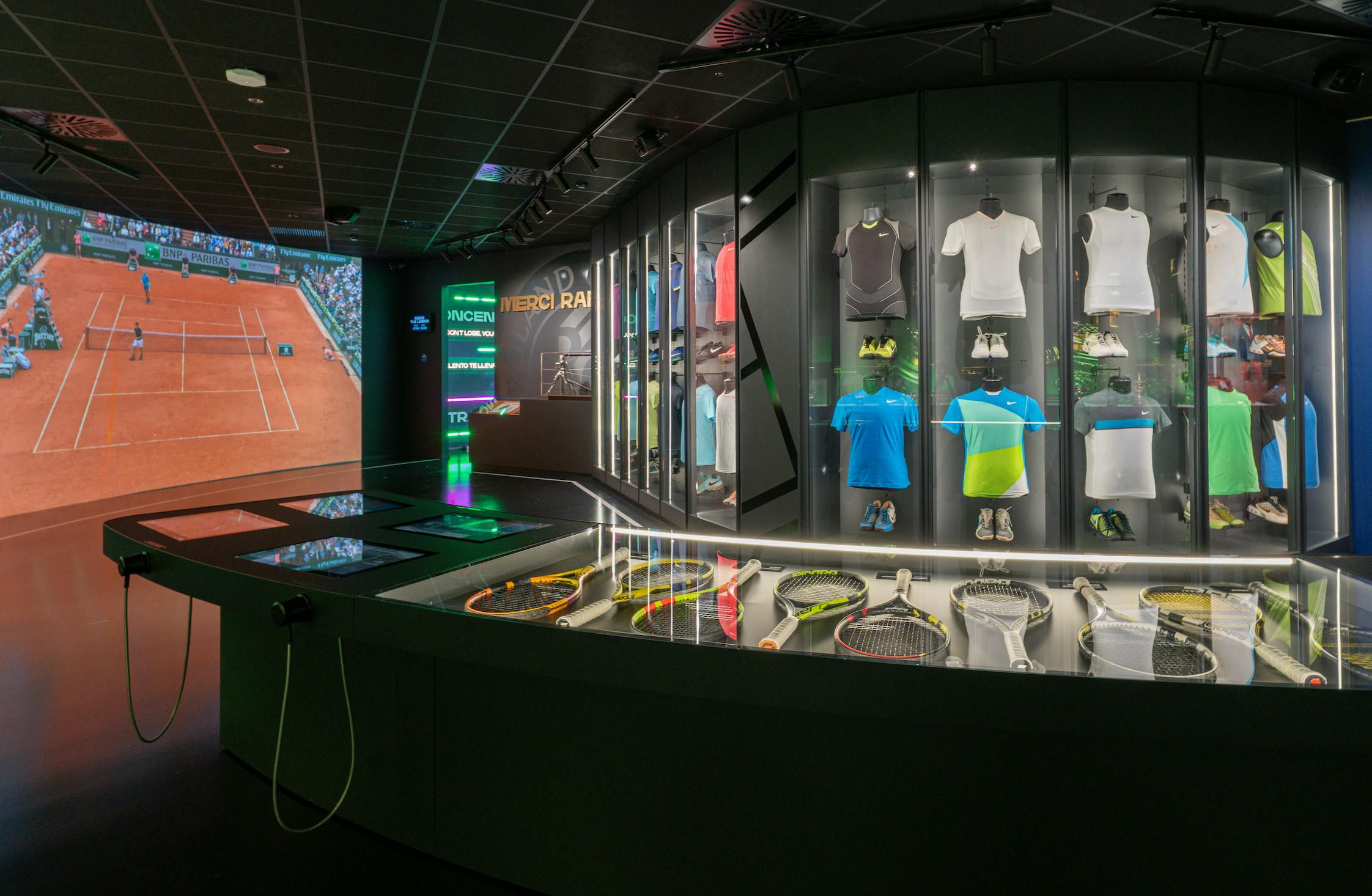 Una zona de exposición temática de tenis con ropa, raquetas y una pantalla que muestra un partido de tierra batida con iluminación interior.