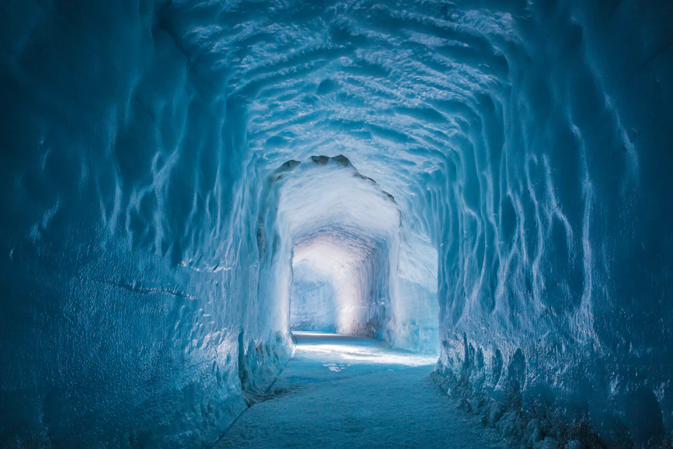 A tunnel made entirely of ice with a smooth, arched ceiling and walls, illuminated by light at the far end.