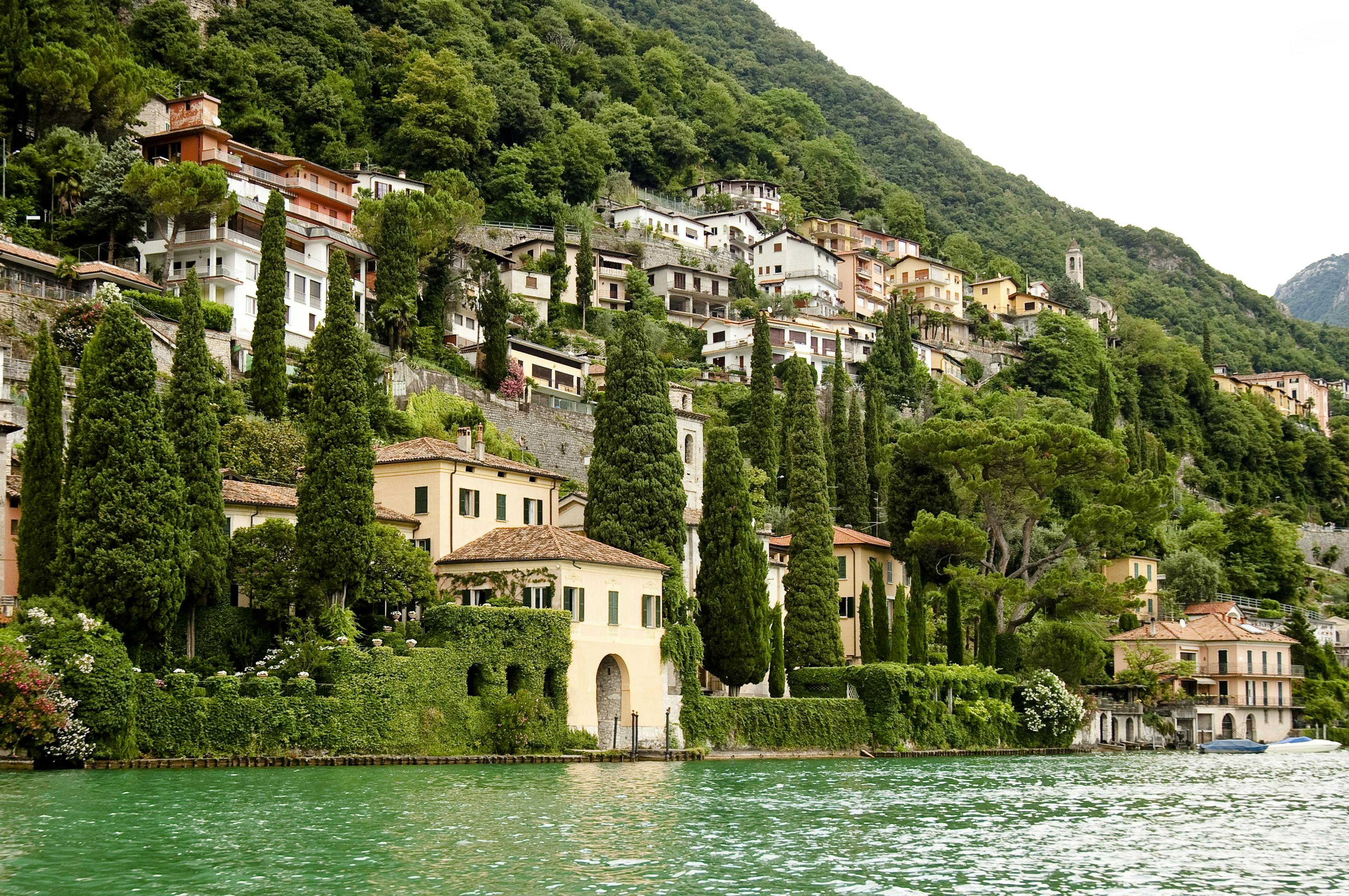 Hillside village with colorful houses, tall cypress trees, and lush greenery along a lake’s shoreline.