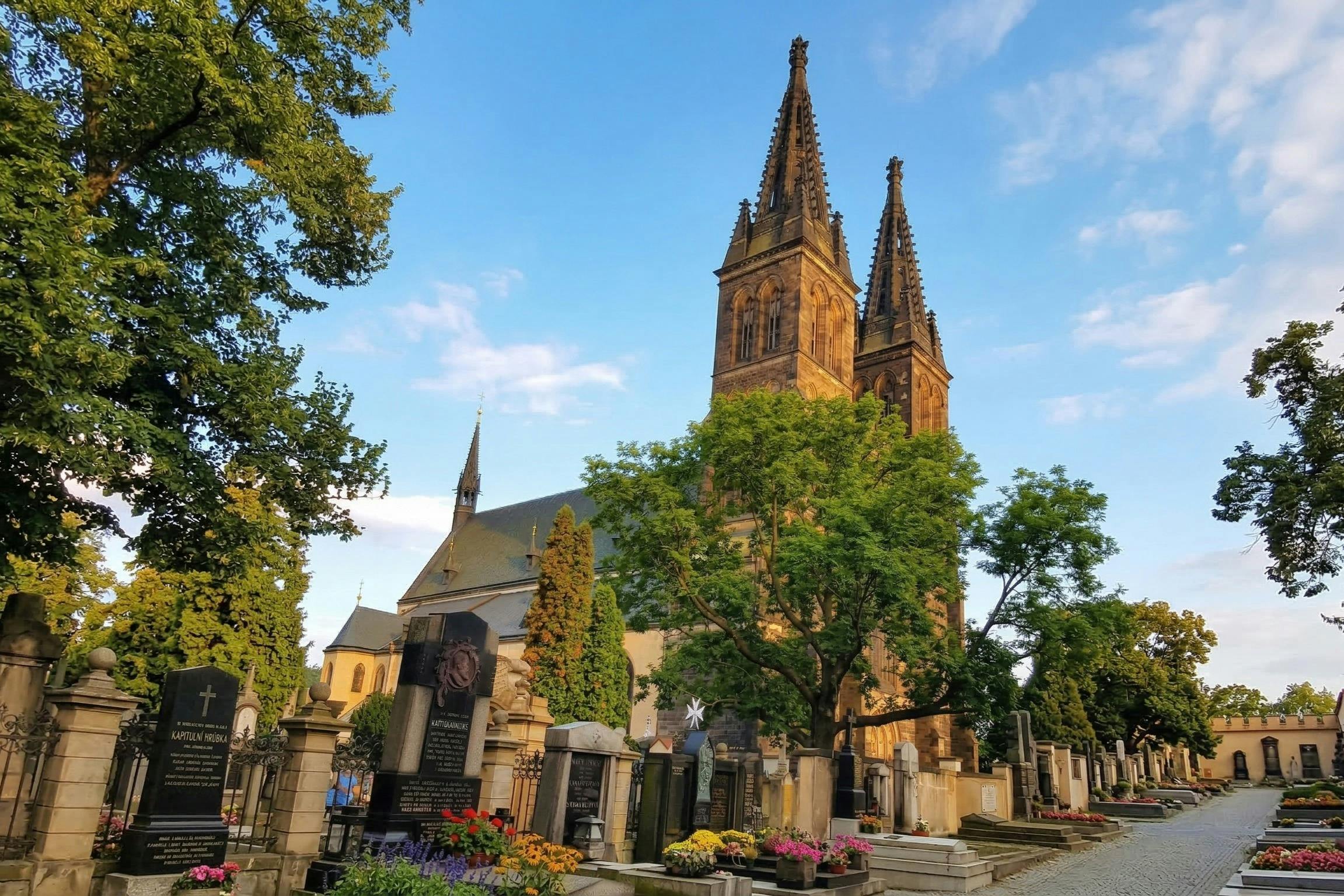 A church with two tall spires behind a cemetery with colorful flowers, gravestones, and trees under a blue sky.