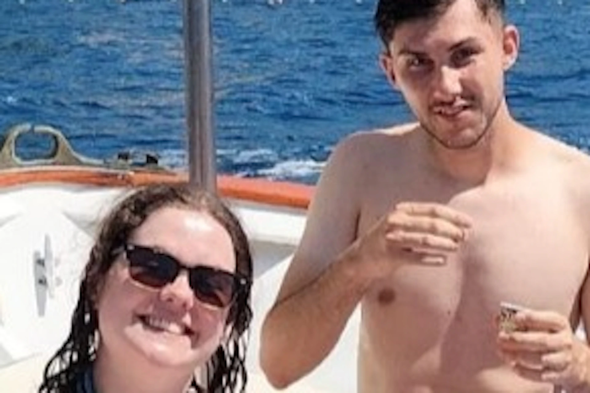 Three people on a boat enjoying drinks with a coastal village and hillside in the background under a clear sky.