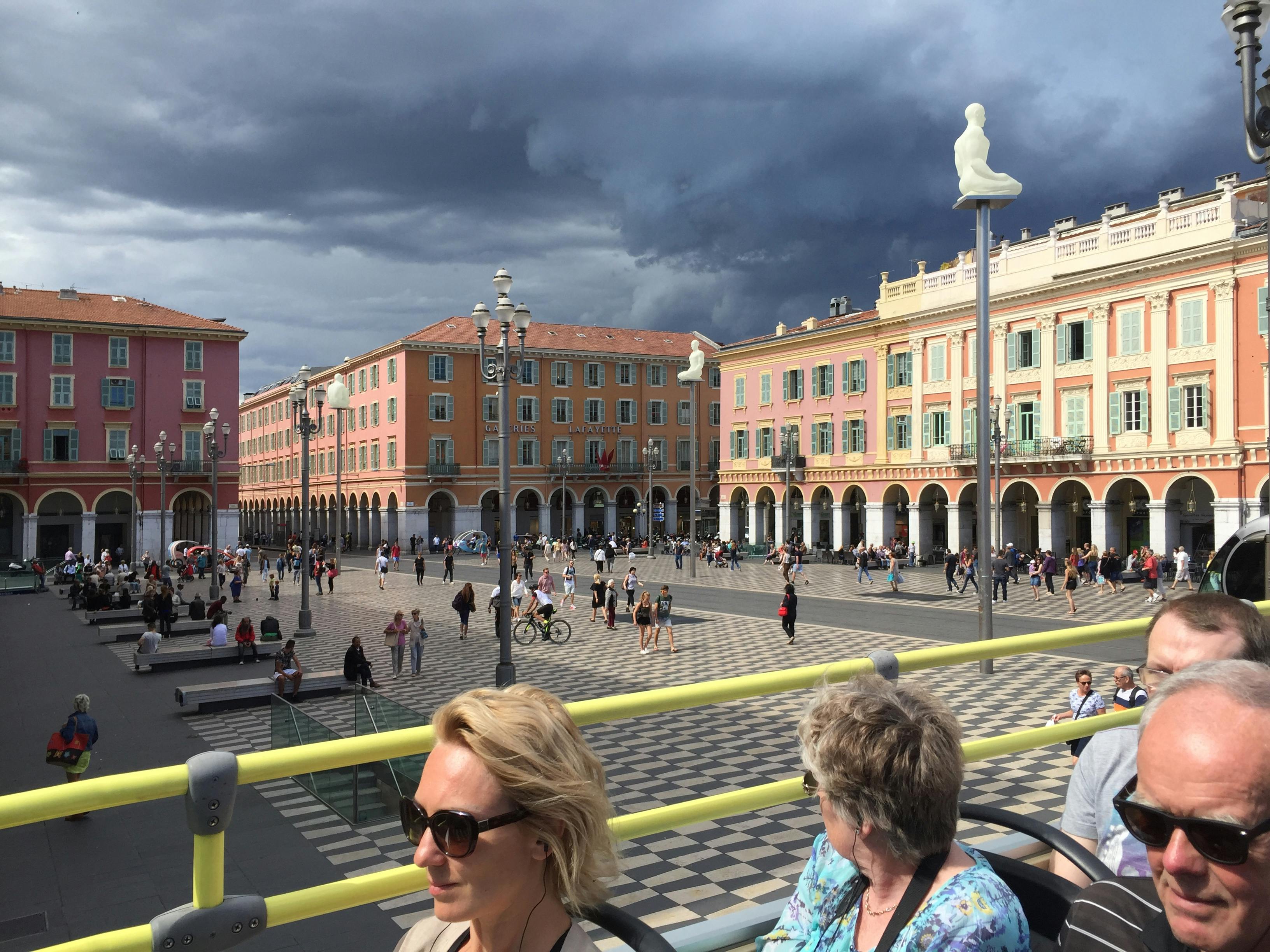 La gent camina i socialitza en una gran plaça envoltada d'edificis de colors sota un cel ennuvolat; dues dones s'asseuen al primer pla.