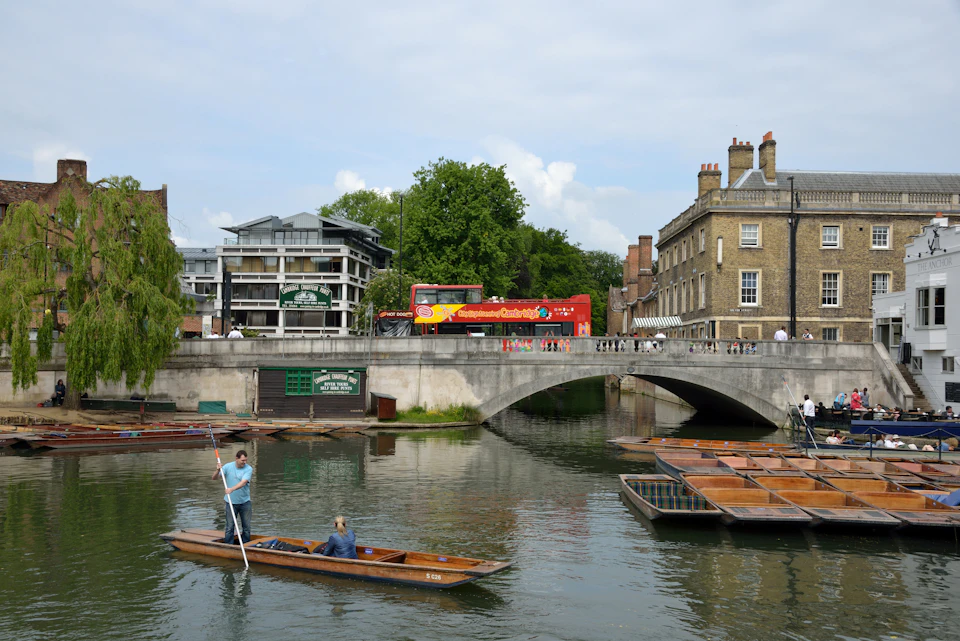 A person punts a boat on a river near a bridge with a red double-decker bus and buildings in the background.