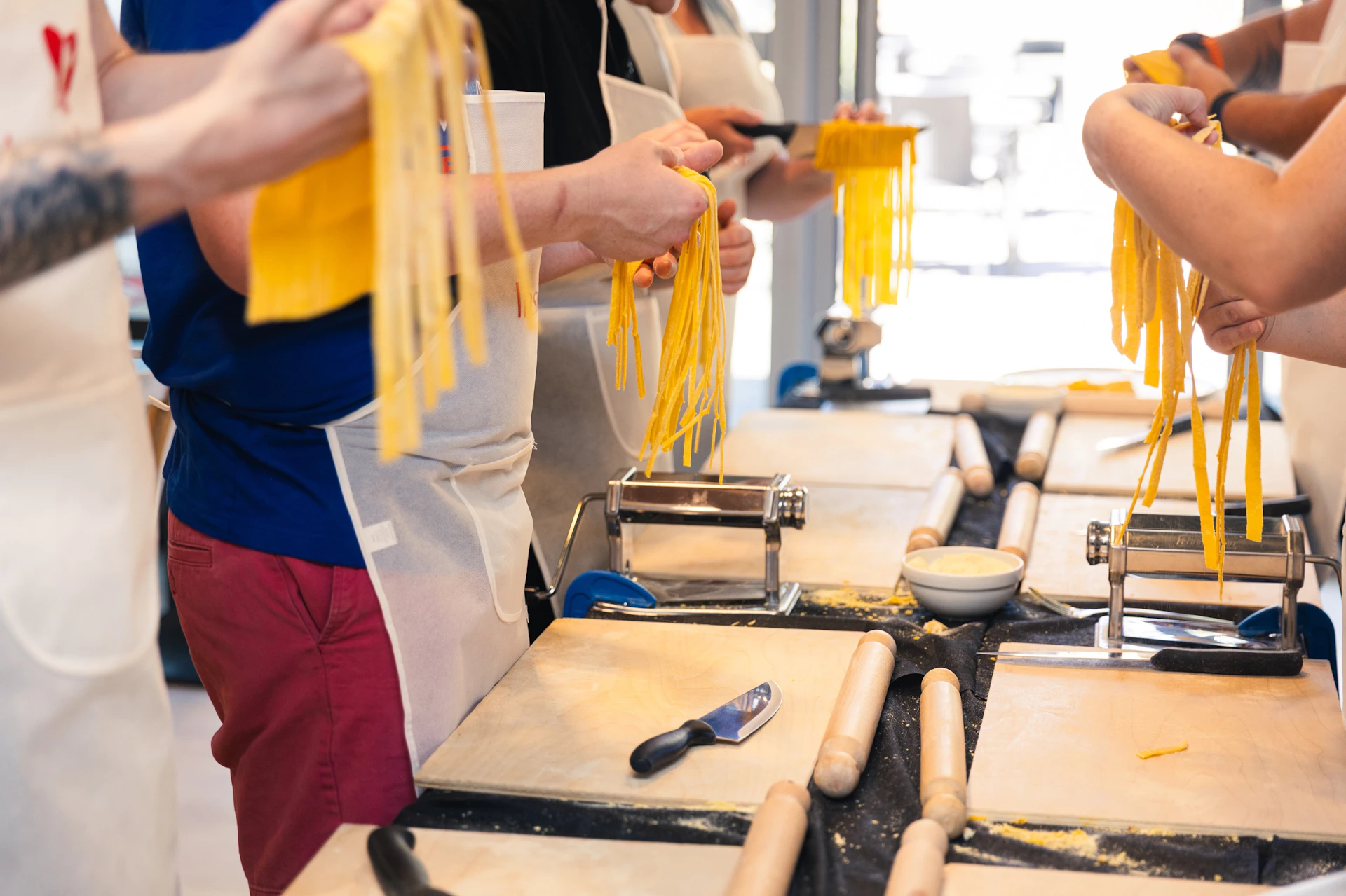 People in aprons making pasta with rollers and cutting boards on a table.