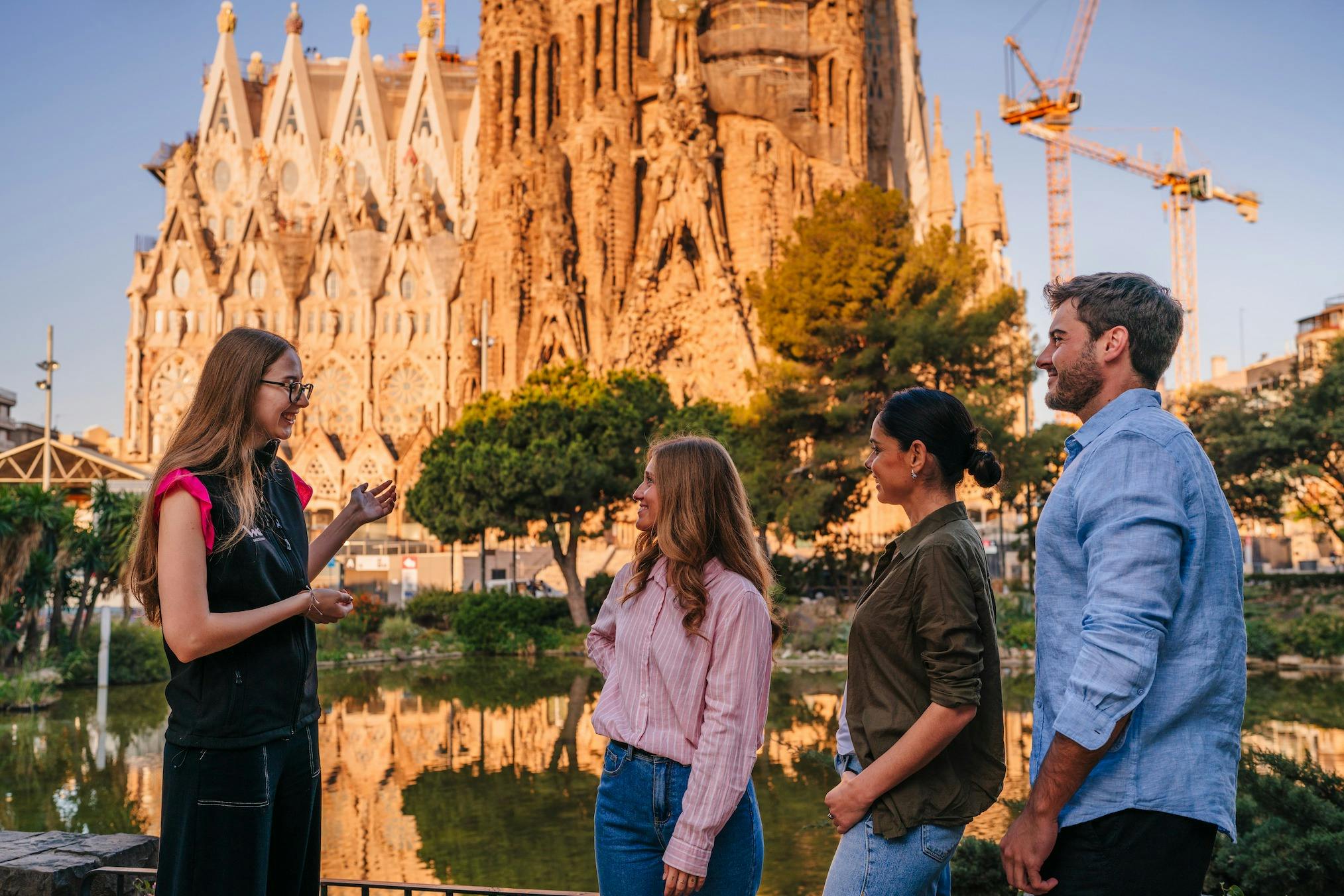 Three people stand outdoors, looking towards a large ornate building surrounded by trees and cranes in the background.